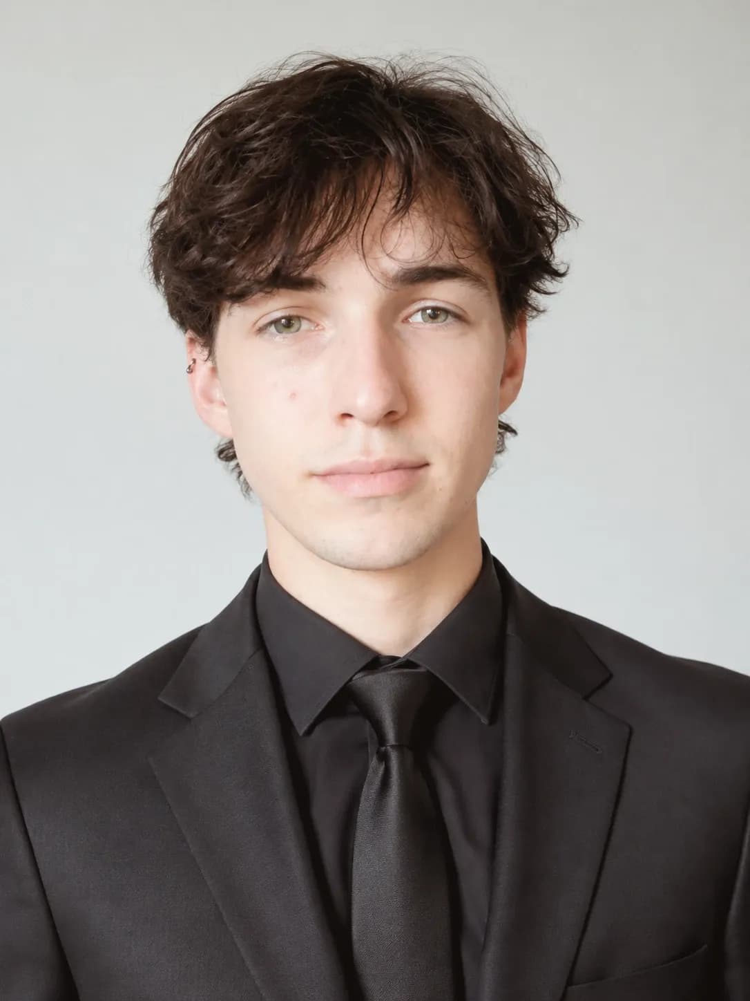 Headshot of Ryan Starrett, a young man with dark tousled hair wearing a black suit, shirt and tie against a light grey background